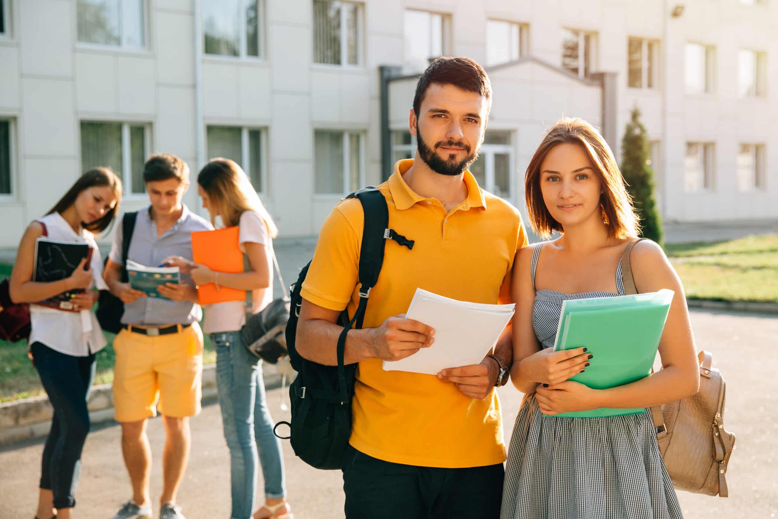 O que é graduação: dois alunos na área externa de uma faculdade com mochila nas costas e segurando material de estudo, ao fundo três alunos conversam entre si.