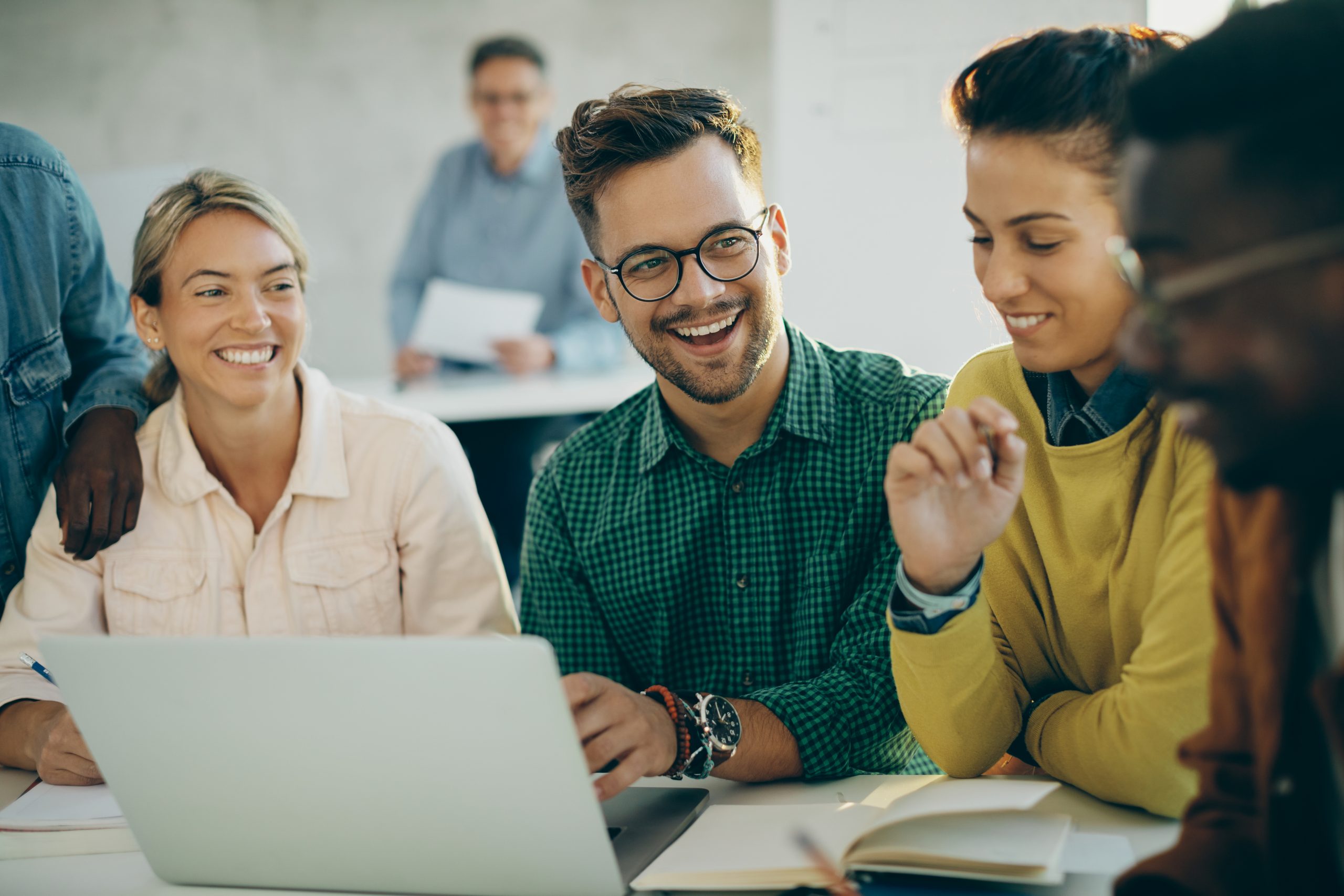 Grupo de estudantes sentados em uma mesa em frente a um notebook rindo e fazendo anotações.