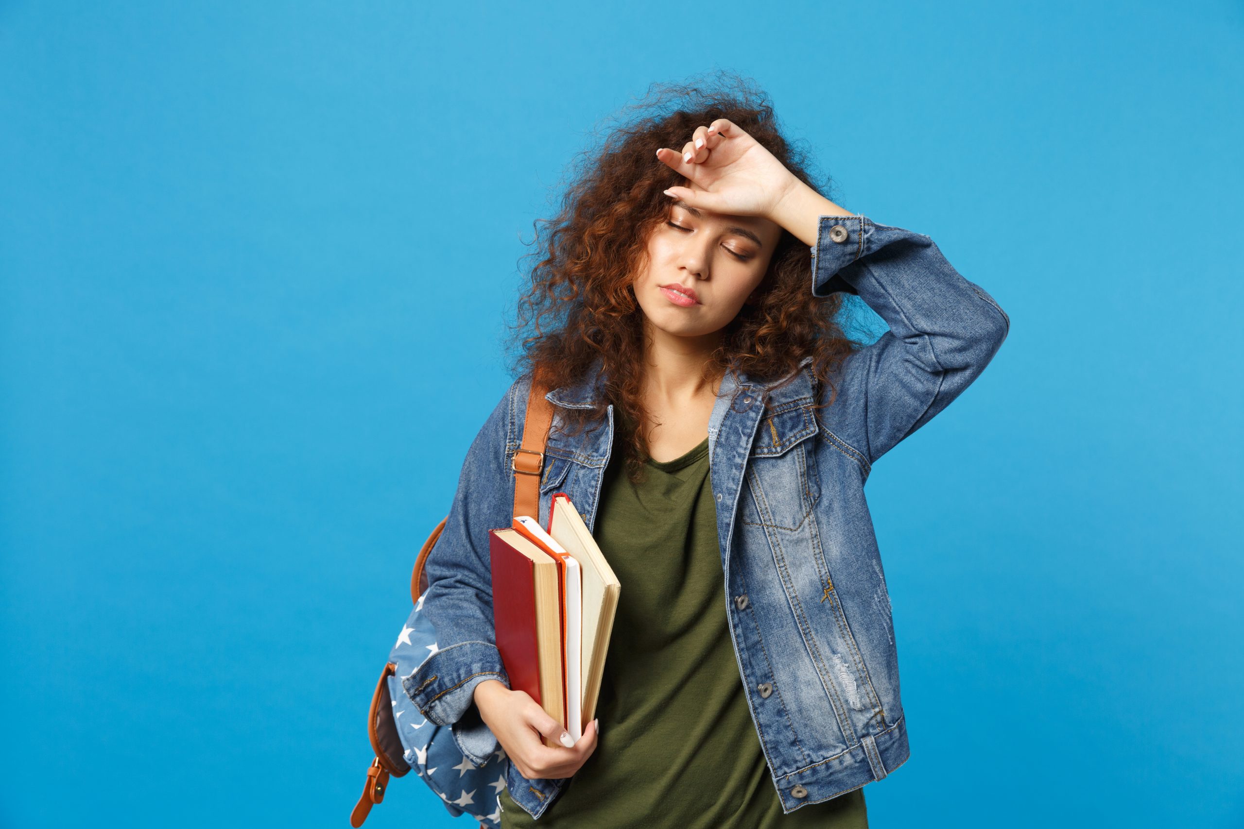 Estudante de aparência cansada com uma mão na testa e a outra segurando alguns livros.