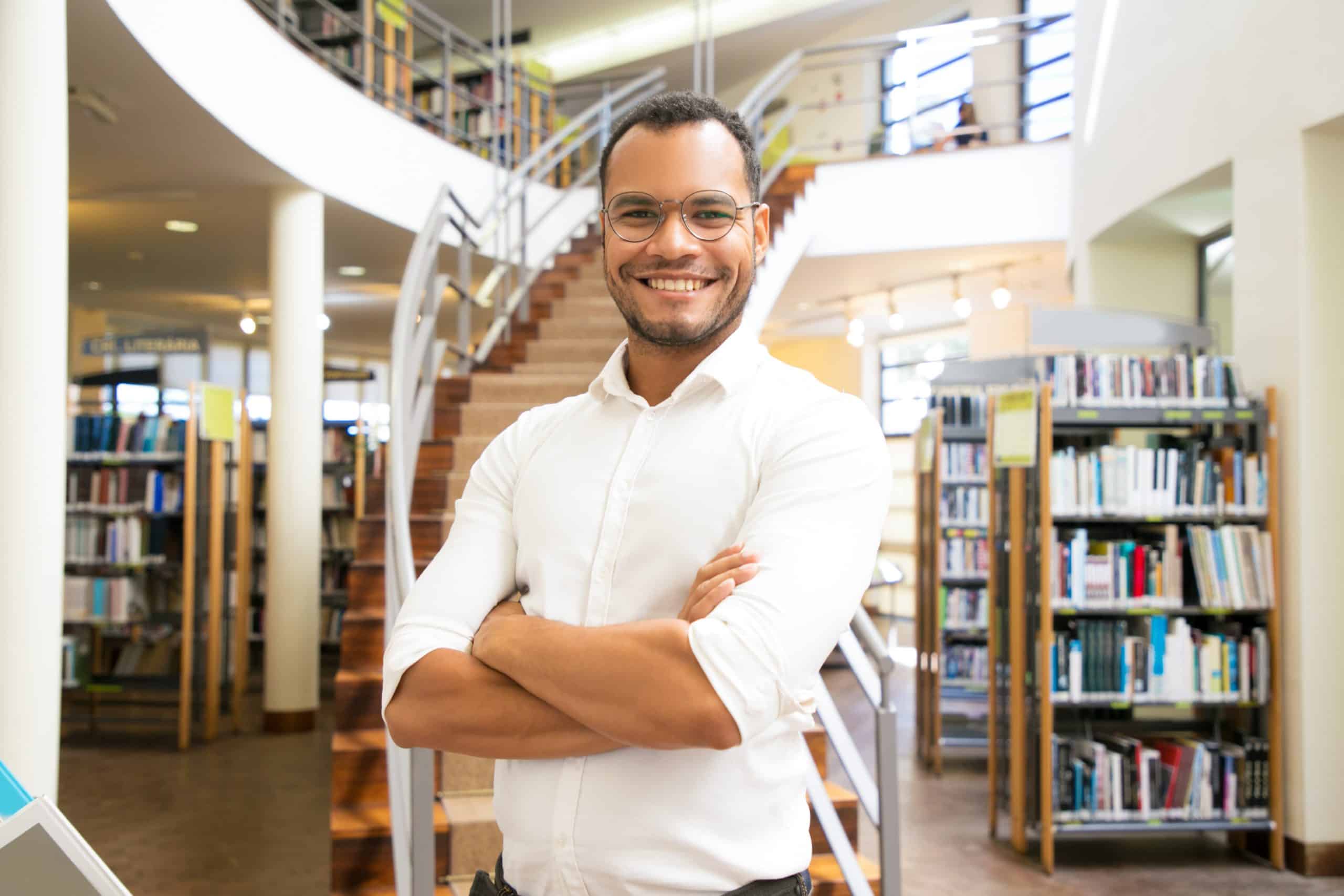 Um homem sorrindo parado e de braços cruzados no meio de uma biblioteca.