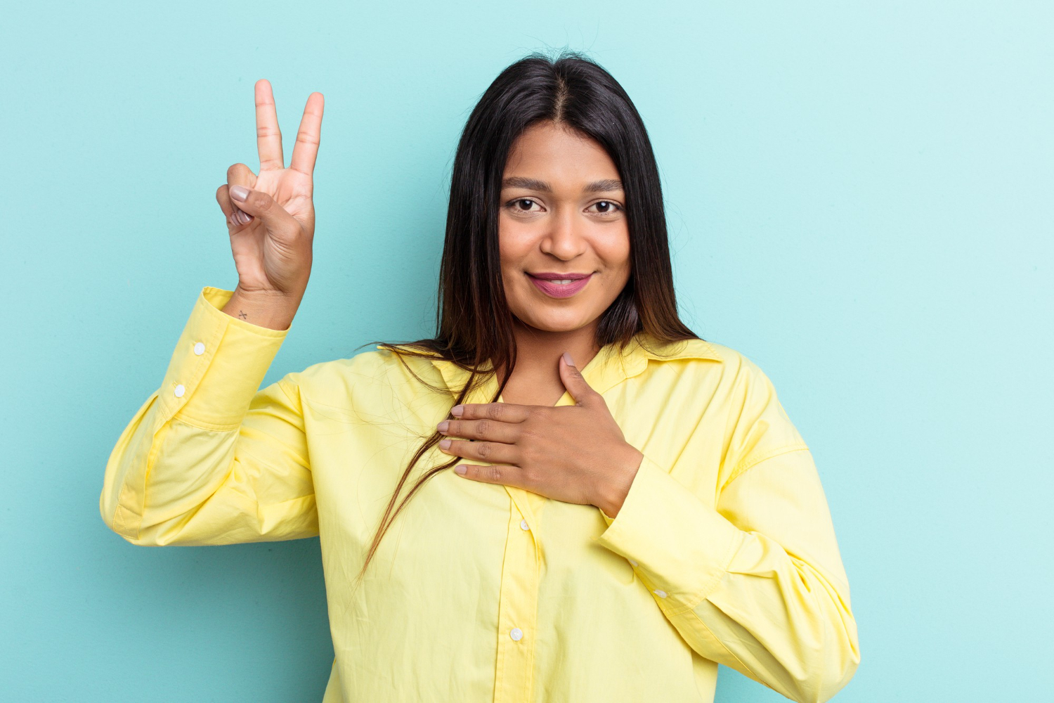 Mulher sorridente demonstrando sinais da linguagem brasileira de sinais com mão na cabeça e outra fazendo o símbolo de paz, contra fundo de cor azul.