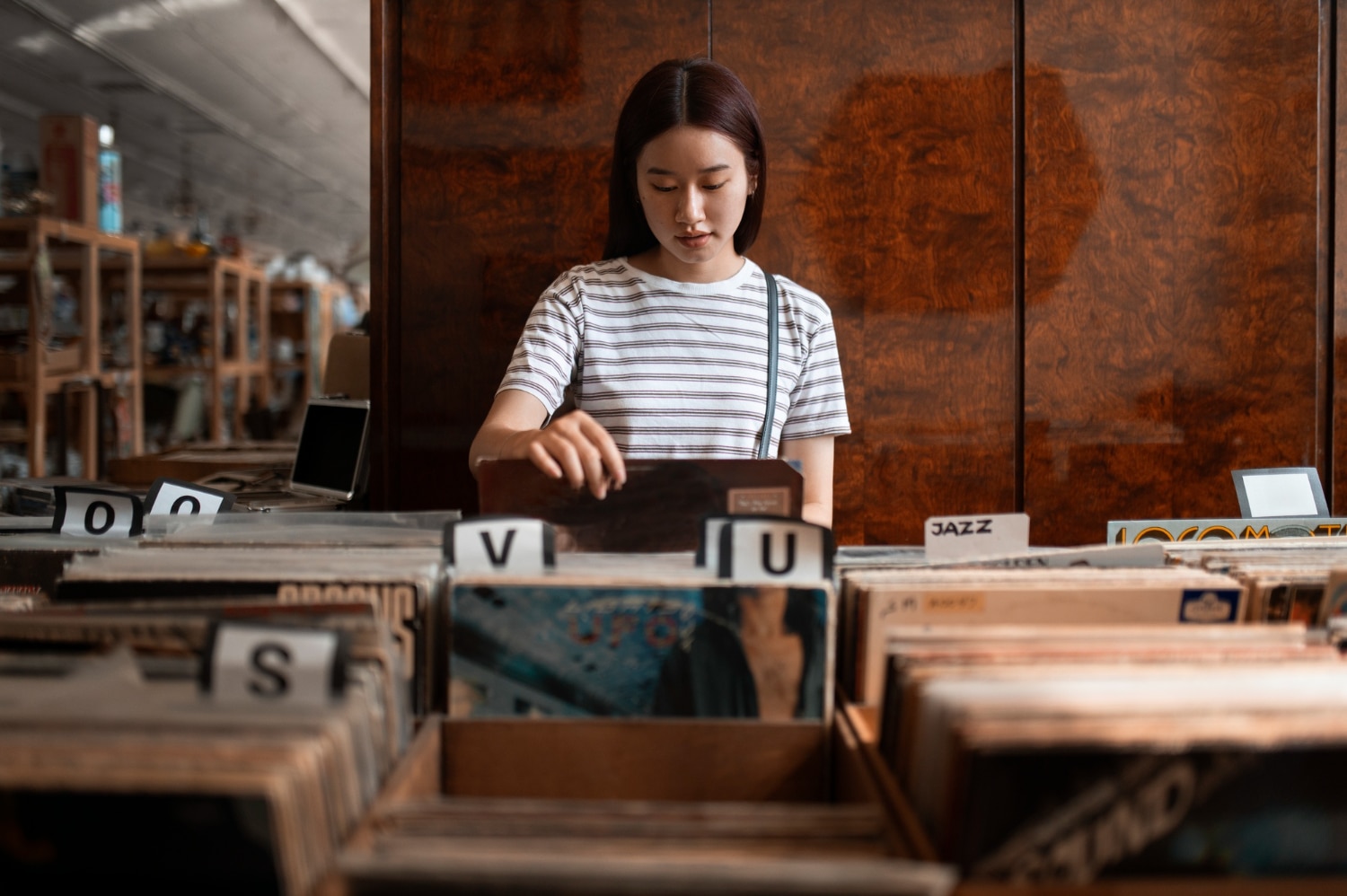 Jovem mulher pesquisando em uma loja de discos ou livraria, realizando pesquisa bibliografica com livros e álbuns ao redor.