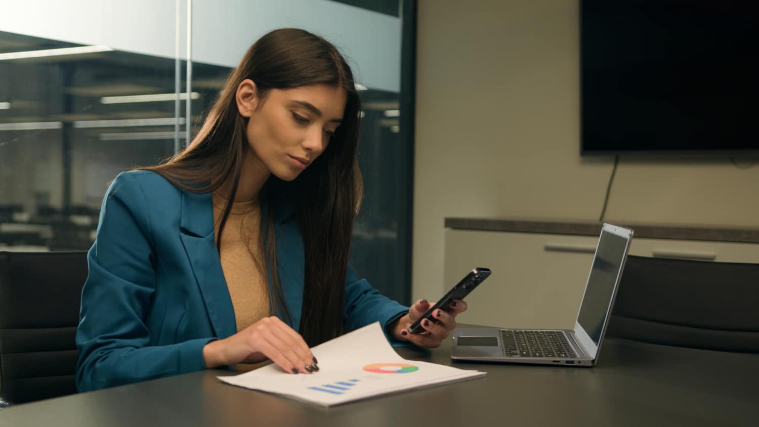 Mulher de negócios analisando salário de um analista de marketing com celular e laptop em uma sala de reunião moderna.